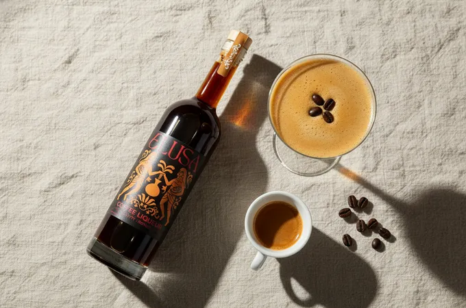 Flat-lay of an Elusa Coffee Liqueur bottle laid on its side on cream linen, beside an espresso martini coupe, espresso cup, and scattered coffee beans